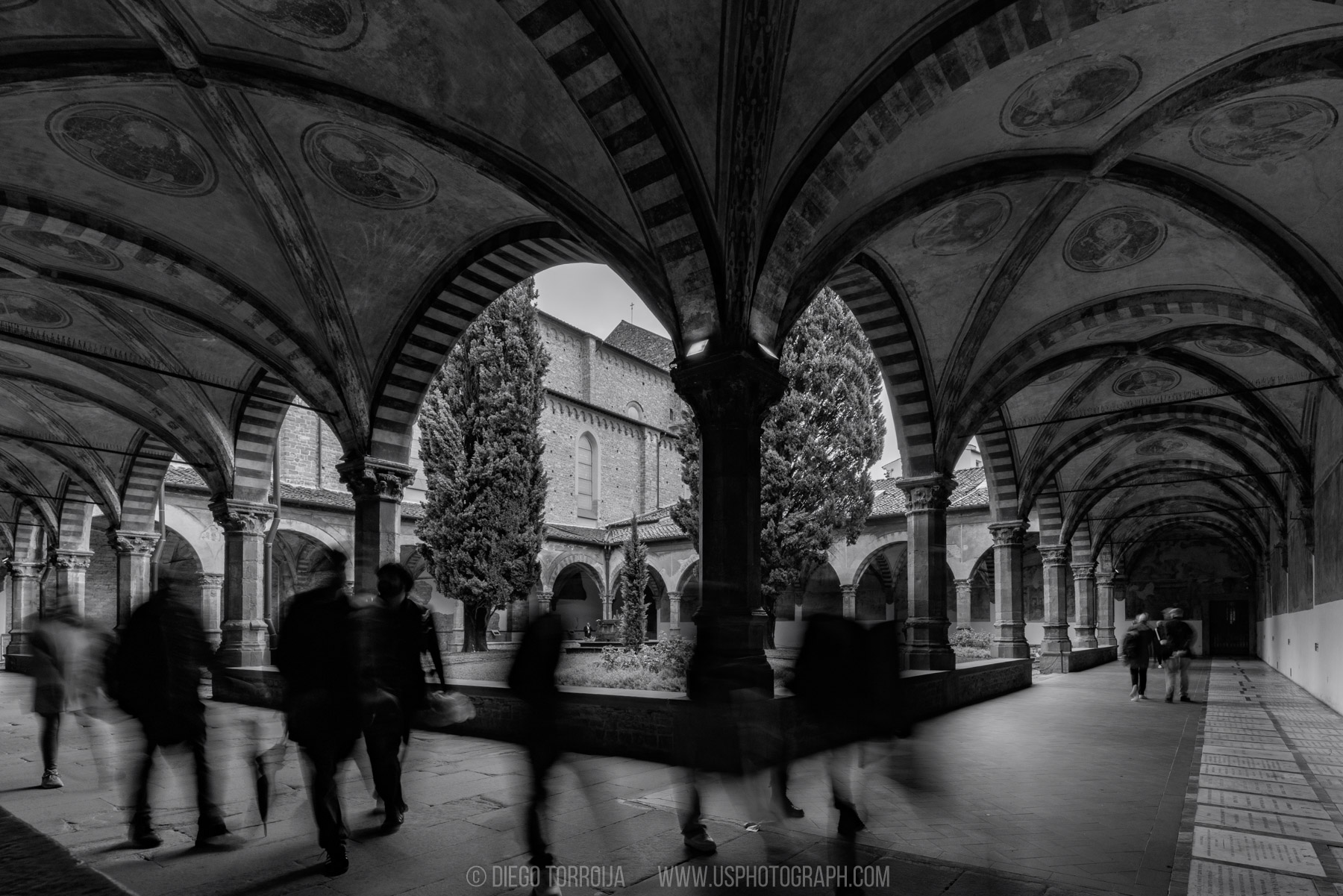 Visitors in motion blur walking through a vaulted cloister with cypress trees in Florence, long exposure photo
