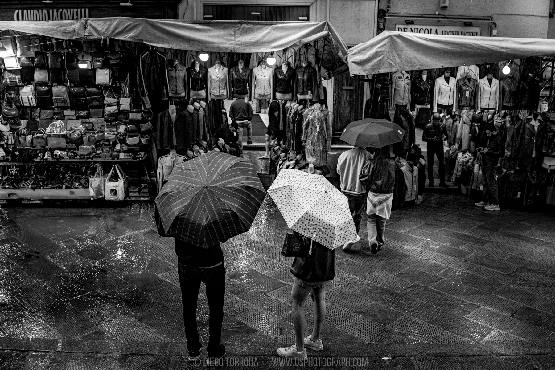 Two people with umbrellas outside leather market stalls on a rainy day in Florence, Italy