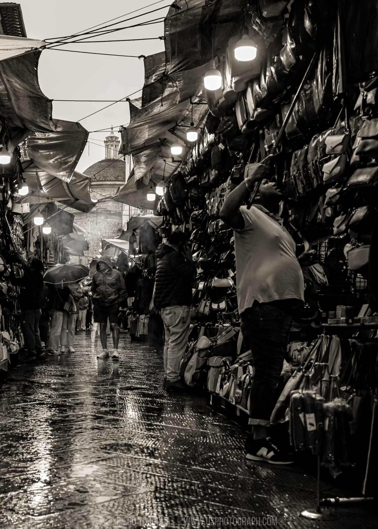Vendor reaching up to leather bags under market lights with a church dome behind, Florence street photography