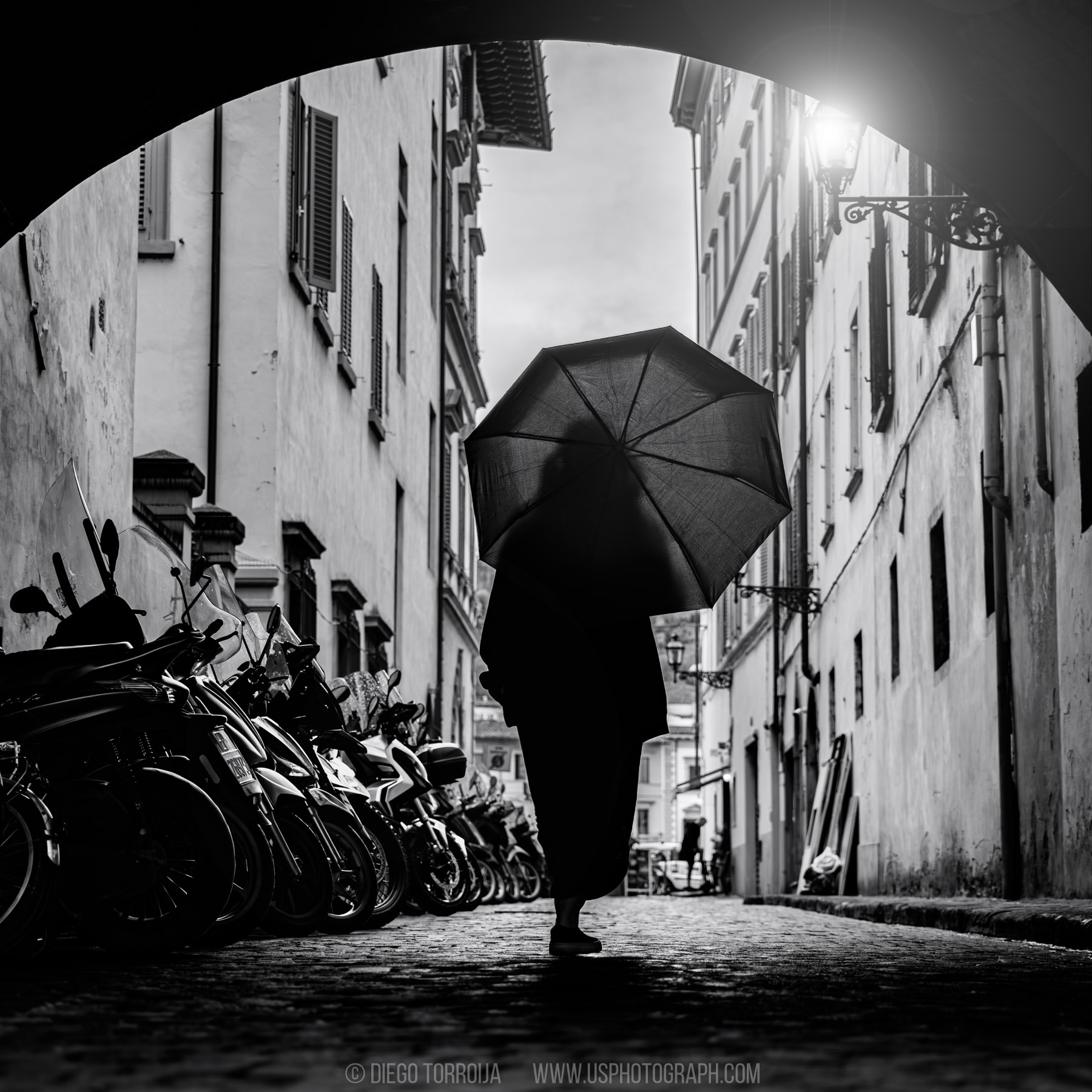 Silhouette of a person with an umbrella walking under an archway past parked scooters in Florence, Italy